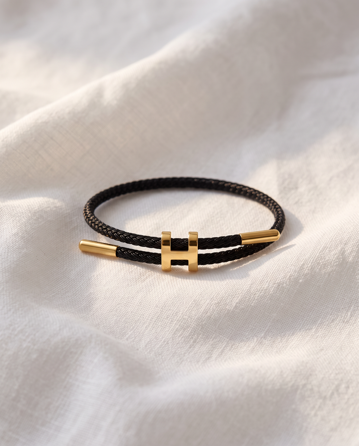 Hills Braid black cord bracelet with gold H centerpiece resting on white linen fabric