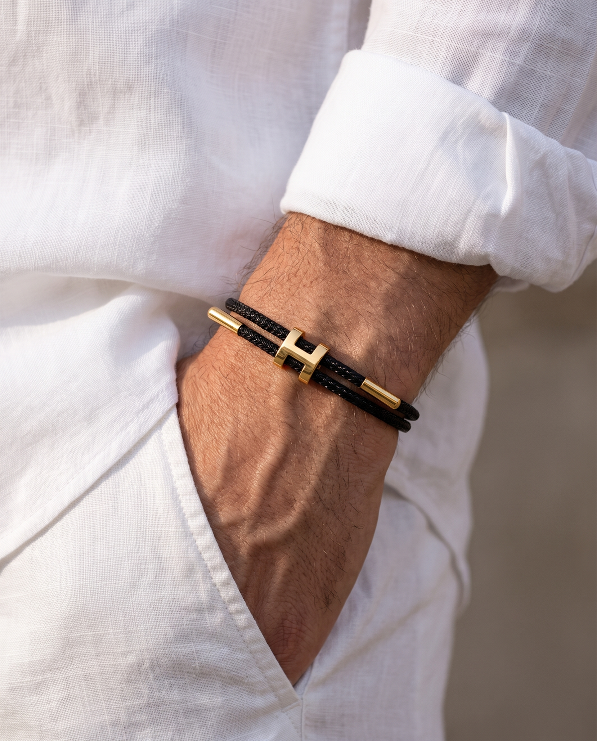 Man in a white linen shirt wearing the Hills Braid black cord bracelet with gold H centerpiece, hand in his white linen trouser pocket